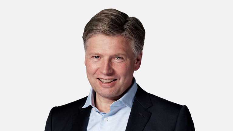 A studio-style portrait of a smiling Sven Smit wearing a dark suit jacket, light blue shirt, and white pocket square against a plain light background.