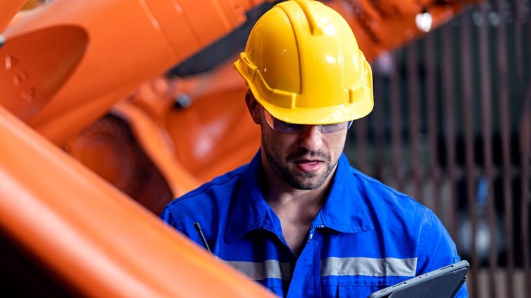 An engineer in a blue jumpsuit and yellow helmet inspects an orange steel welding robot while looking down at a tablet.