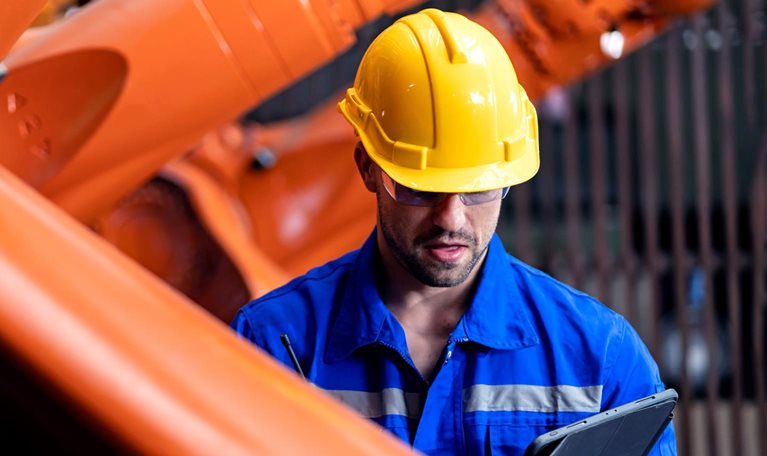 An engineer in a blue jumpsuit and yellow helmet inspects an orange steel welding robot while looking down at a tablet.