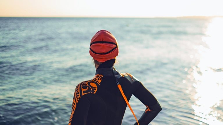A triatalete wearing a wetsuit and swim cap stands facing a calm ocean. Her back is to the viewer as she takes in the setting sun.