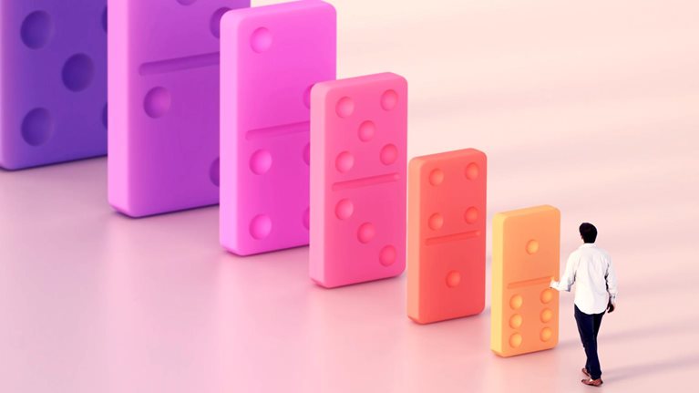 A person approaches a row of increasingly large, colorful dominoes, suggesting a cascading impact.