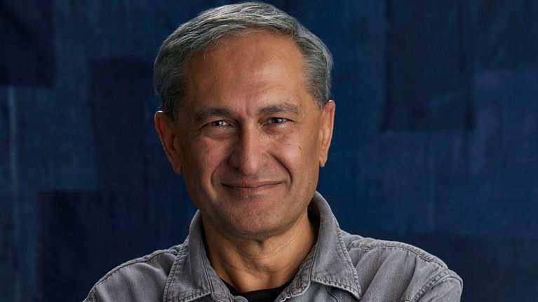 A portrait of a Harmit Singh with short hair and a slight smile, looking directly at the camera. He is wearing a gray button-up shirt and is set against a dark blue textured background.