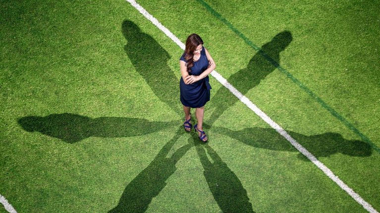 Overhead view of a woman in a dark blue dress standing with her arms crossed on a green sports field, surrounded by six elongated human-shaped shadows. White and dark green field lines cross the grass around her.