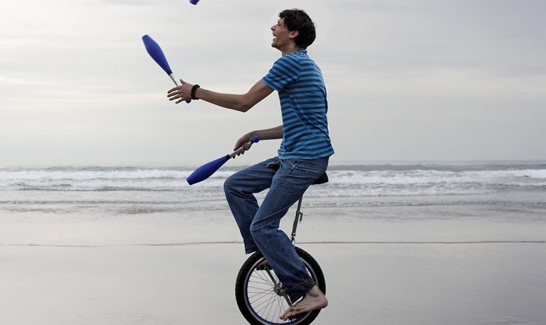 Man sitting on a unicycle while juggling pins at the beach