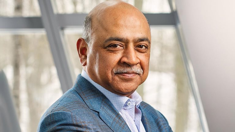 Arvind Krishna with a neatly trimmed mustache, wearing a blue blazer over a light blue collared shirt. He is standing indoors in front of large windows, looking calmly at the camera with a composed, professional expression.
