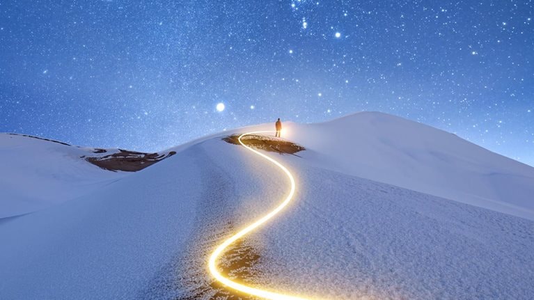 A solitary figure stands upon a snow-covered peak against a starry night sky. A zig-zagging light trail marks the path taken by the figure to reach the summit.