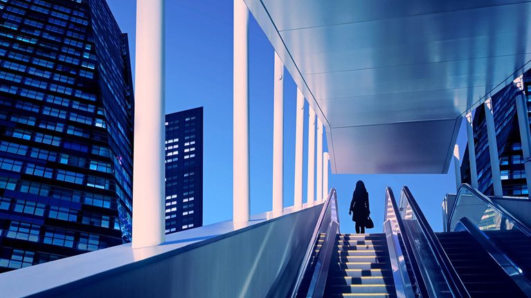 Early evening at modern business district with silhouette of businesswoman on top of a moving escalator.