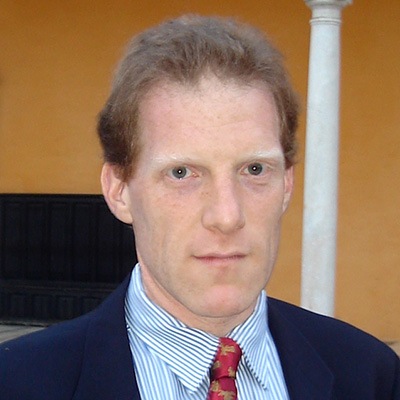 David Greenberg standing in front of a terracotta wall with Doric columns. He is wearing a navy blue suit jacket and a red patterned tie.