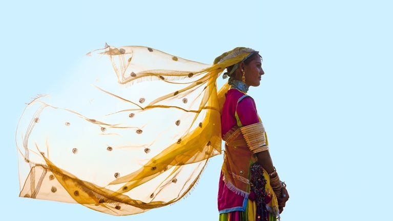A woman from India stands tall and proud in a vibrant saree, her yellow veil fluttering in the wind against a backdrop of a clear blue sky.