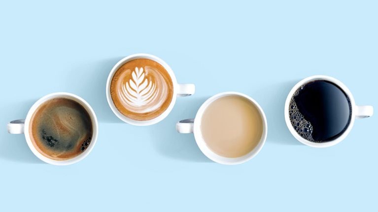 Overhead view of cups with different types of coffee. One of the cups sits higher up in the frame with a foam flourish on top.