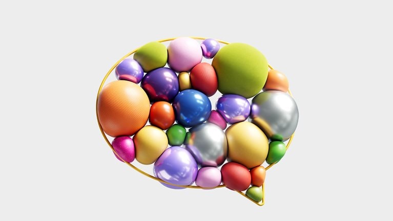 A golden outline of a speech bubble is filled with a jumble of colorful, balloon-like spheres. The spheres vary in size, color, and texture, and the image is set against a light-gray background.