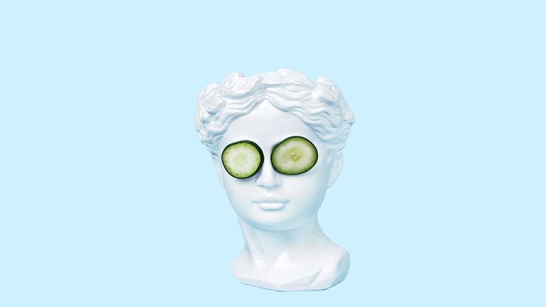 A bust of a young woman, inspired by ancient Greek art, wearing cucumber slices on her eyes and set against a light blue backdrop.