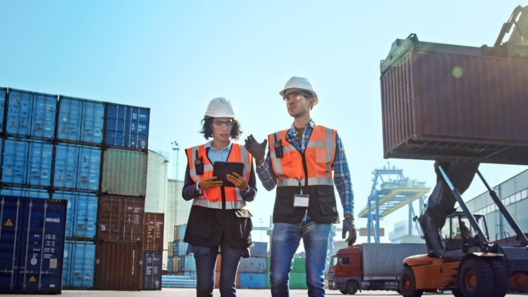 Female industrial engineer with tablet computer and male foreman worker in hard hats and safety vests walk in container terminal