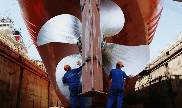 The image captures workers performing maintenance on a large ship's propeller and rudder in a dry dock. This process involves essential inspections and upkeep.