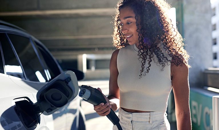 Young woman holding electric plug by car at charging station