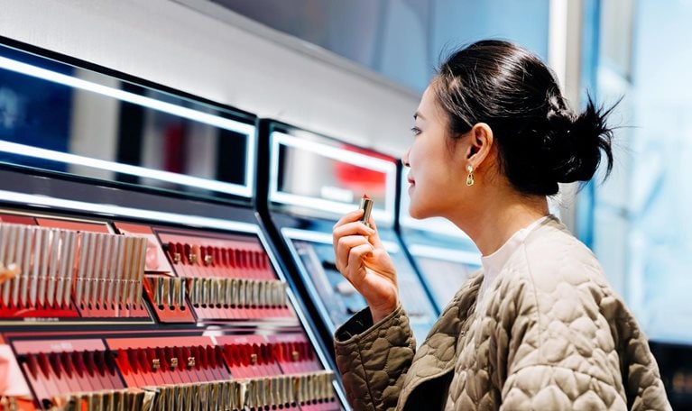 A woman applying lipstick at a makeup counter. The counter has rows of lipsticks in various shades of red and pink, and is lit by bright lights.