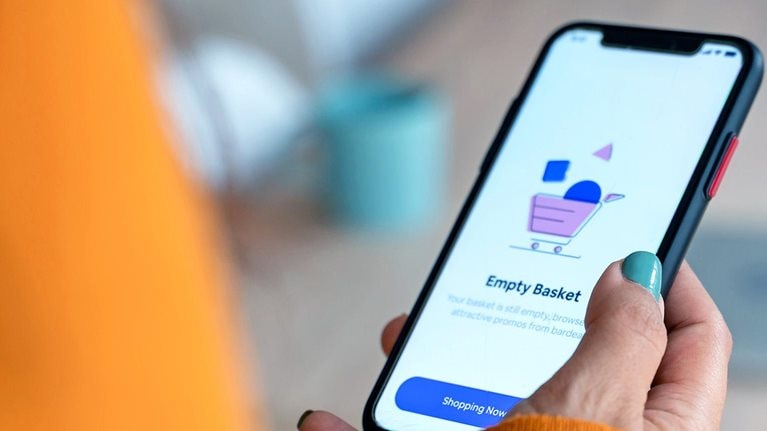 Person in an orange sweater holding a smartphone displaying a shopping app screen that says “Empty Basket”