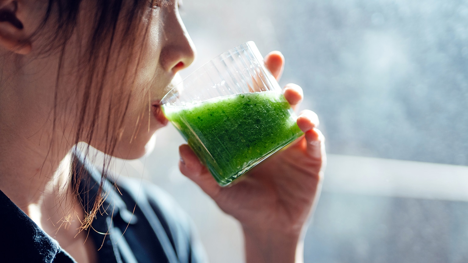 A close-up of a woman enjoying a bright green smoothie from a transparent glass.