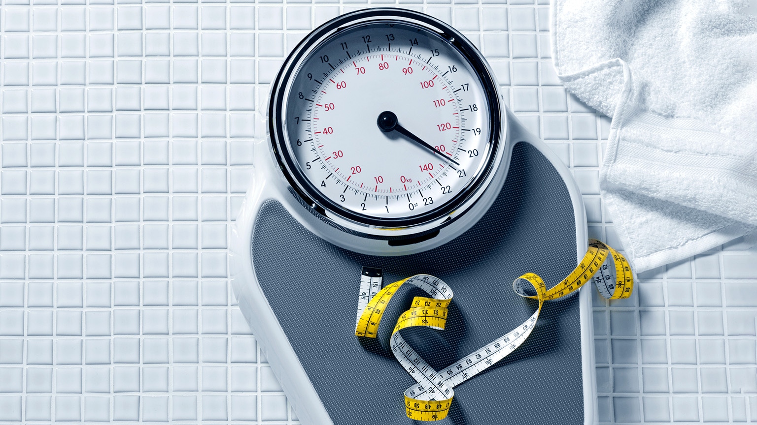 A bathroom scale with a large, round face sits on a tiled floor, next to a folded white towel. A yellow measuring tape is coiled on the scale's platform.