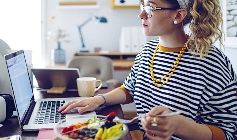 A woman wearing glasses and a striped shirt is seated at a desk, looking at her laptop and eating a salad.