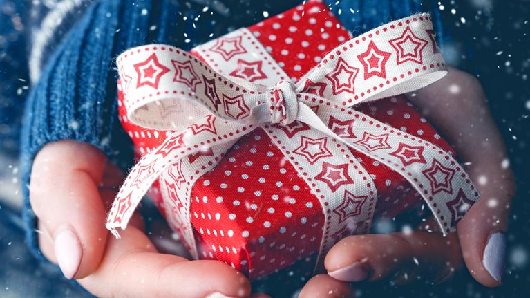 Close-up image of hands holding a small red gift box with a festive white bow with stars on it, with snow falling in the background.