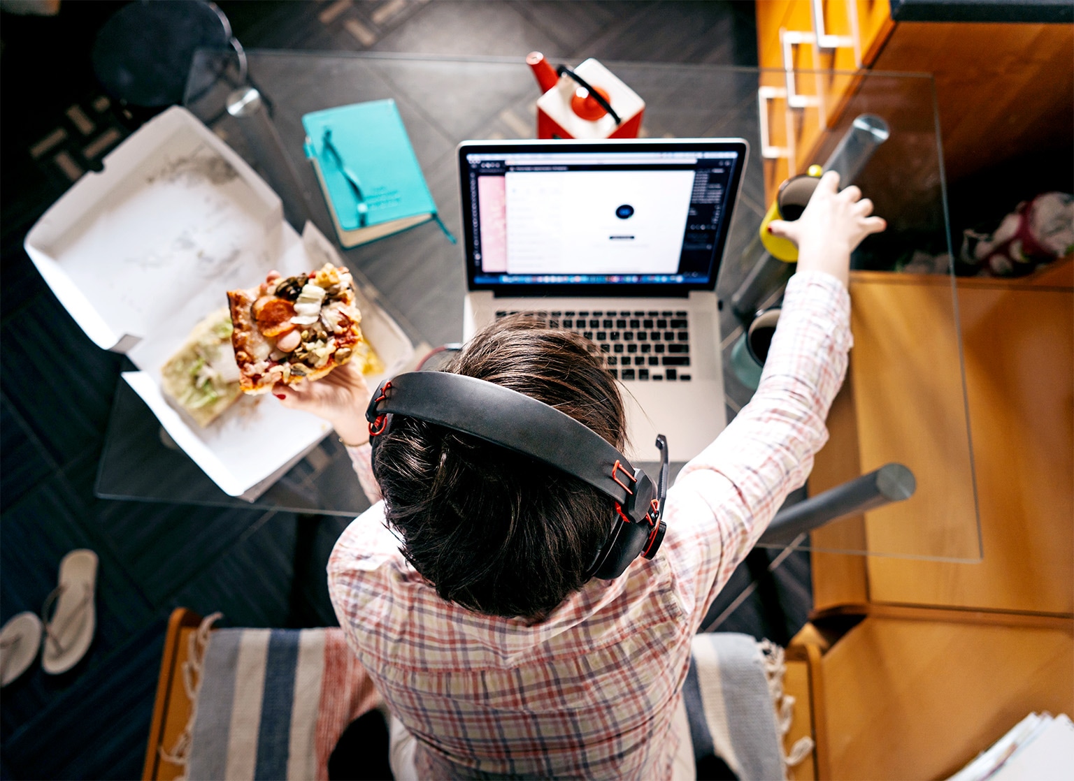 A top down view of a person sitting at a desk in front of a computer eating delivery pizza.