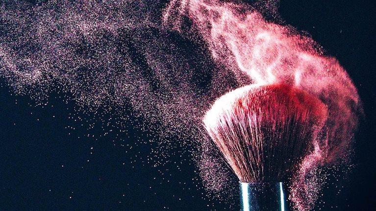 A makeup brush with blush particles artistically being shaken off