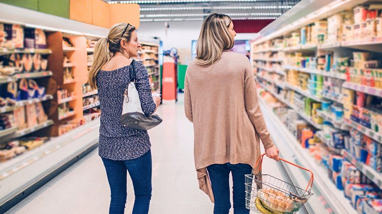 Two women walking down a grocery store aisle with a shopping basket