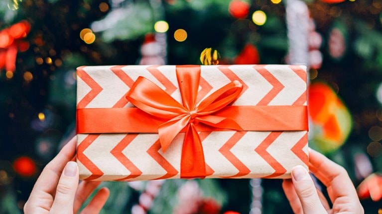 A pair of hands holding a wrapped gift box decorated with a red chevron pattern and tied with a bright red ribbon. In the background, colorful lights and ornaments create a festive, holiday atmosphere.