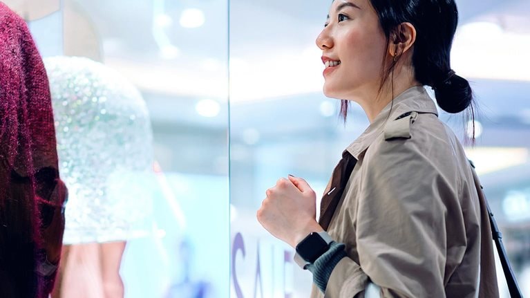 Asian woman carrying a shopping bag, standing outside a boutique looking at window display in a shopping mall