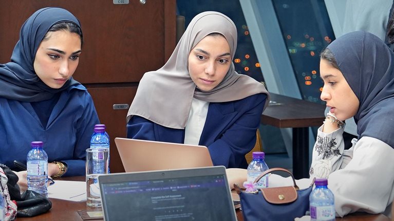 In a modern room overlooking city lights, a group of people sit around a large table with multiple laptops, notebooks, and water bottles. Several women wearing headscarves focus on a laptop while others, including a man with clasped hands, take notes or lean over to observe.