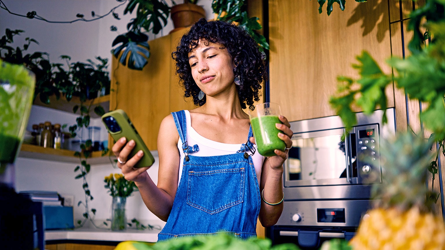 A young woman wearing stylish overalls stands in the middle of a modern kitchen with many plants. She looks for information on her phone while holding a healthy green drink.