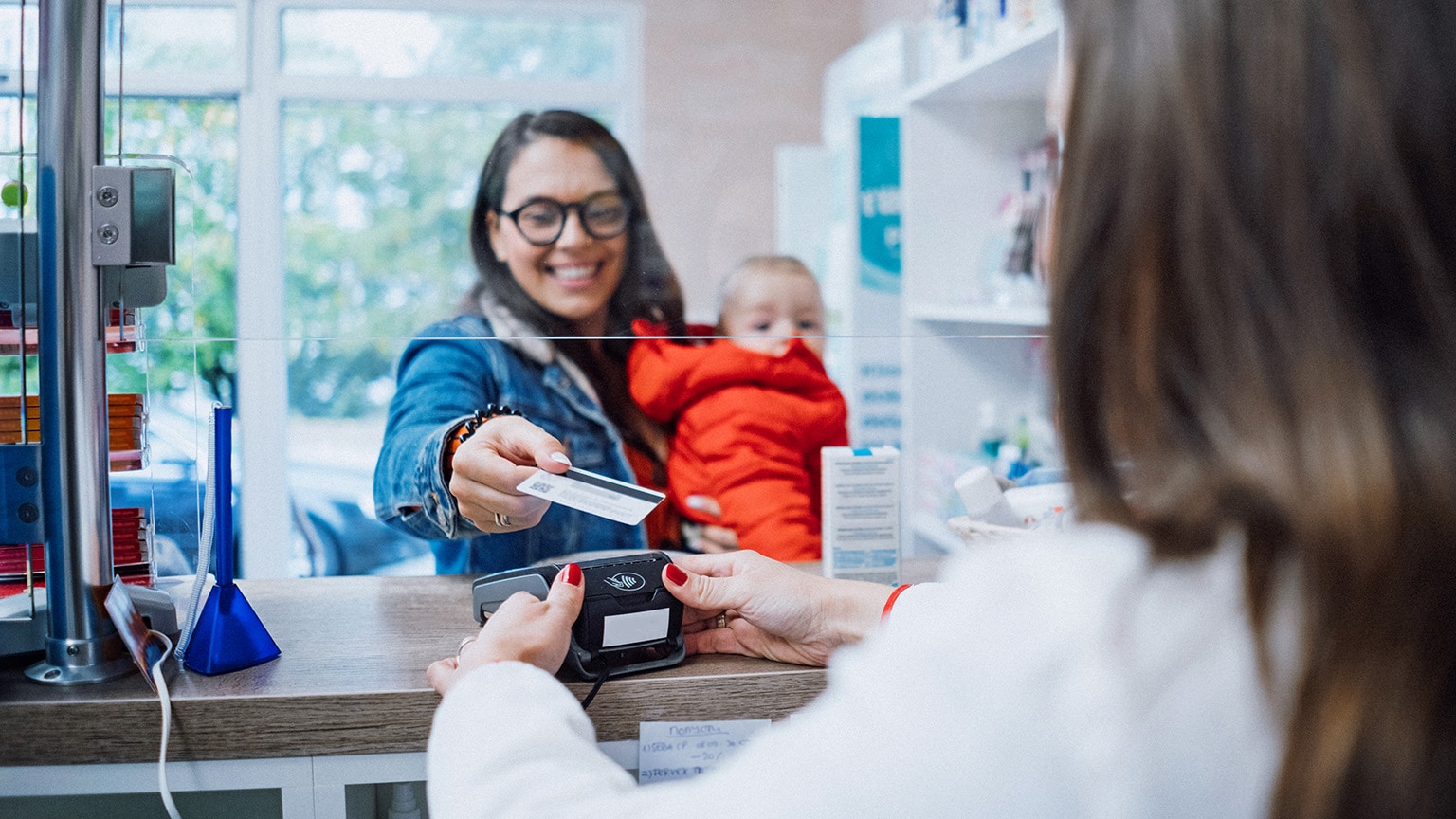 A smiling woman, holding a young child in a red puffy coat with one arm, hands her credit card to a pharmacist.