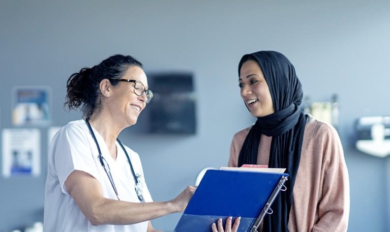 In an examination room with medical equipment behind them, a female patient wearing a black hijab and a pink sweater is greeted by a female doctor dressed in a white coat and stethoscope. The doctor holds a clipboard and smiles warmly at the patient.