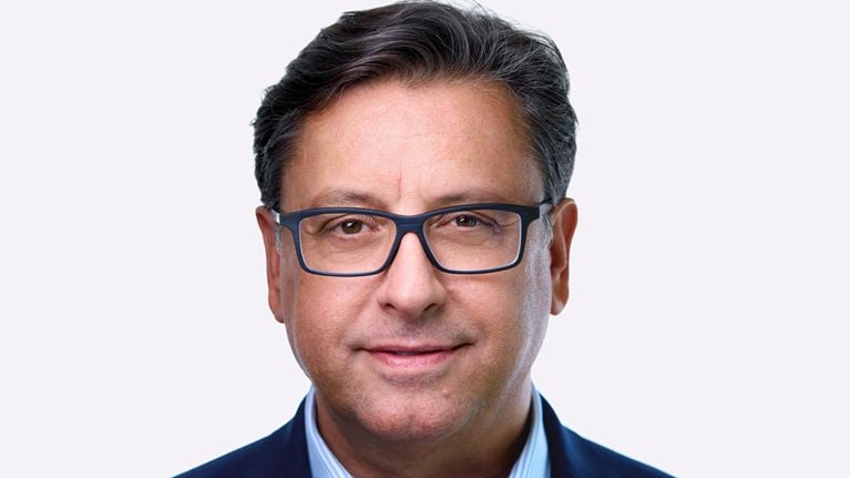 A man named Paul Hudson, sporting dark hair and glasses, can be seen in a slight smile as he faces the camera. He is dressed in a blue suit jacket and a striped blue shirt.