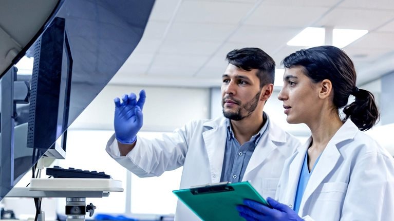 Two laboratory scientists in white coats and blue gloves stand side by side examining a large machine with a monitor. One scientist points to the screen while the other holds a clipboard, both appearing focused on the data or results displayed.