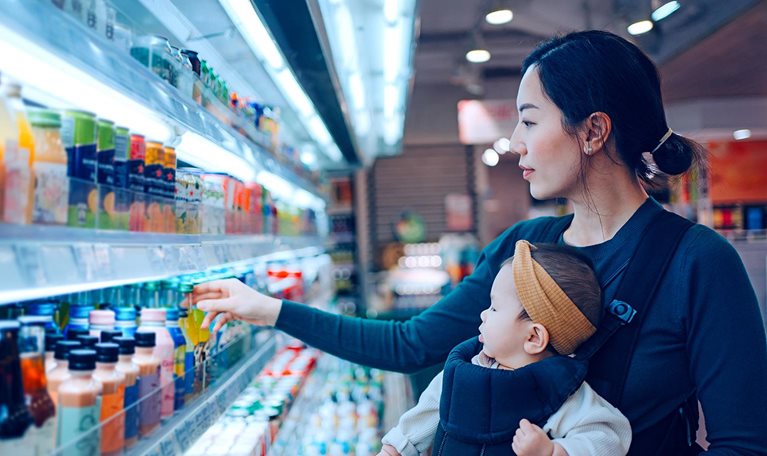 Young Asian mother carrying her baby girl in a baby carrier, doing grocery shopping in supermarket. Choosing fresh organic fruit juice for her family. Healthy eating habits with balanced diet.