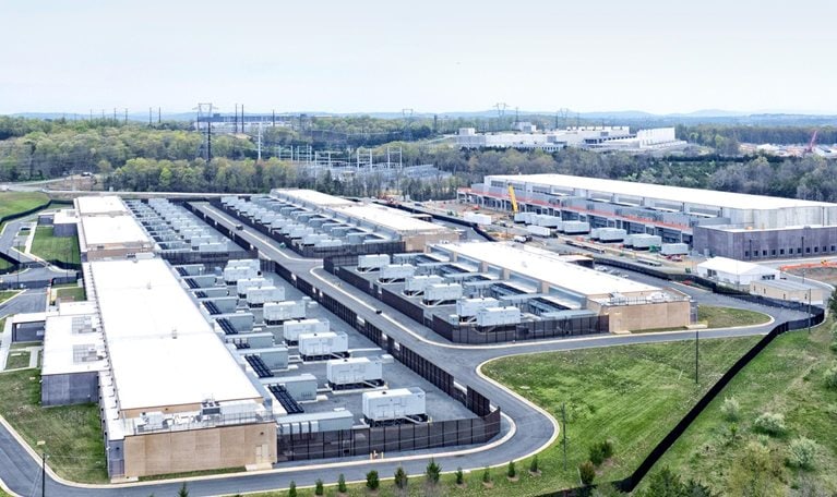 A sprawling data center complex surrounded by greenery and infrastructure. Several rectangular buildings with white roofs are visible, densely packed with cooling units or similar equipment, with at least one building under construction.