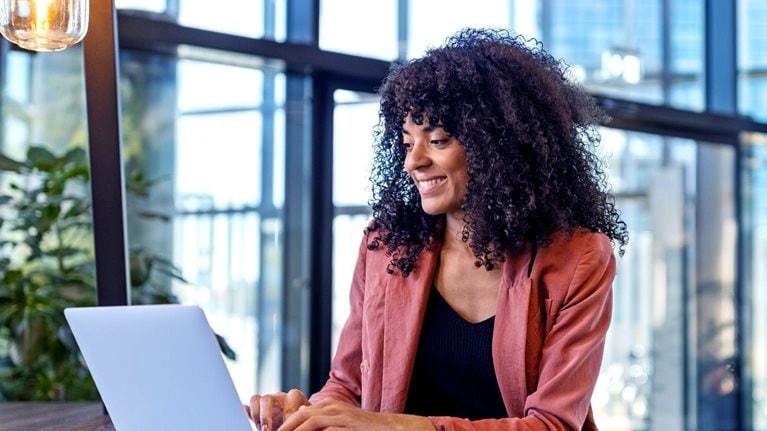 A cheerful young lady is seated at a table in a contemporary and well-lit cafe, typing away on her laptop.