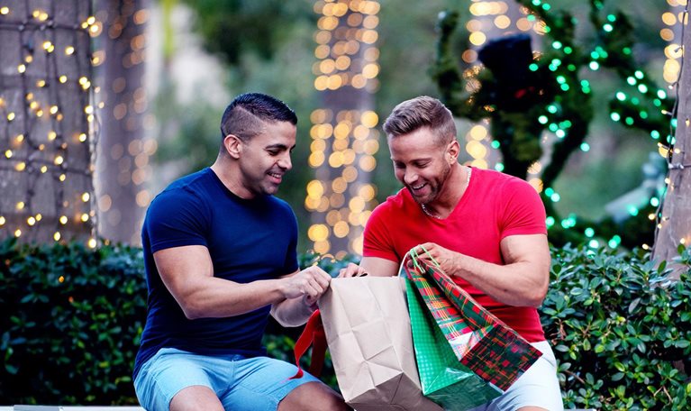 Two men dressed for warmer weather look through their purchases while holding holiday themed shopping bags. String lights adorn the trees behind them.