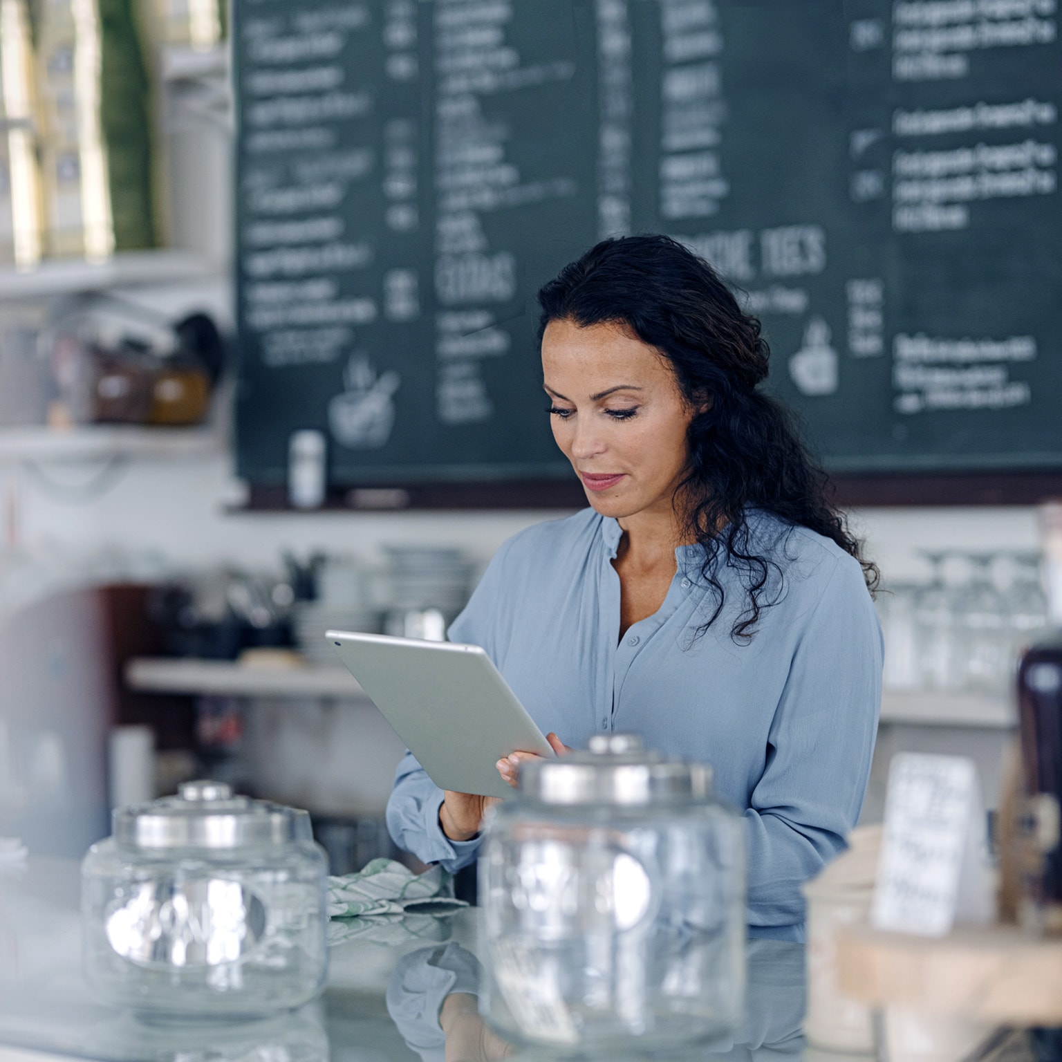 Brunette woman using digital tablet while standing at counter in coffee shop with the menu behind her.