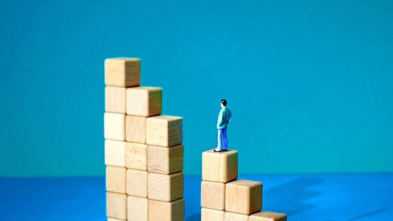 A series of wooden blocks arranged in a stair-like formation, with a gap in the middle where a vertical set of blocks is missing. A tiny man stands, unable to continue forward with his arms hanging at his sides.