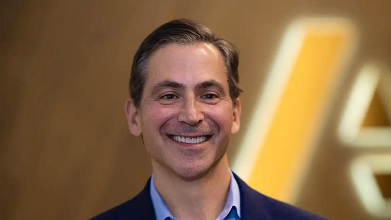 A smiling Charlie Gottdiener dressed in a suit coat and button-up shirt, set against a blurred backdrop featuring the Anaplan logo.