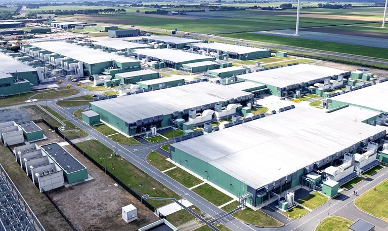 High angle view of a data center surrounded by green fields and wind turbines on the horizon.