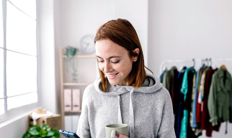 Woman dressed in gray hoodie looking down and phone in one hand, other hand holding ceramic coffee mug.