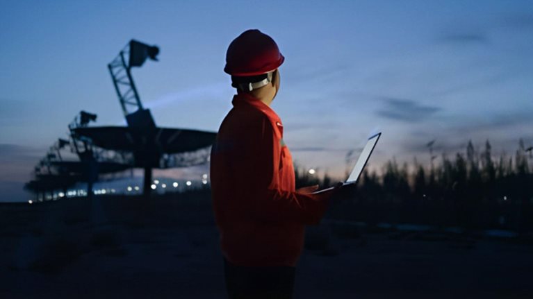 Photograph of a person wearing a red safety helmet and jacket holding a laptop, standing outdoors at dusk near large telecommunications equipment. The scene highlights a work environment with artificial lighting and a darkening sky, suggesting monitoring or inspection activities during low light conditions.