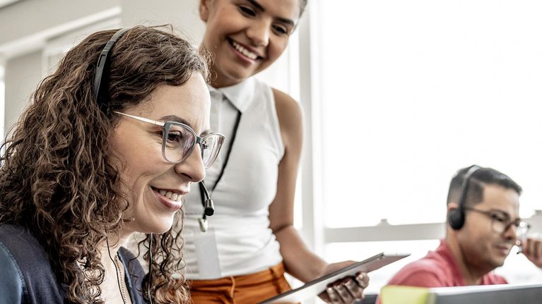 Photograph of a modern office setting with three people engaged in work activities. One person with curly hair sits at a desk, another stands holding a tablet, and a third works on a laptop, highlighting a collaborative and technology-driven work environment.