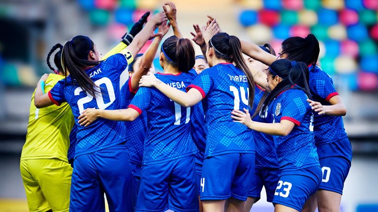 A professional women's soccer team forms a circle with their arms raised and embracing.