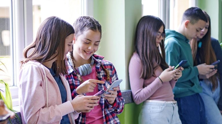 A group of teenagers are standing against a wall, looking at their smartphones. One of the girls in the middle is pointing at her phone, sharing something with another girl beside her.