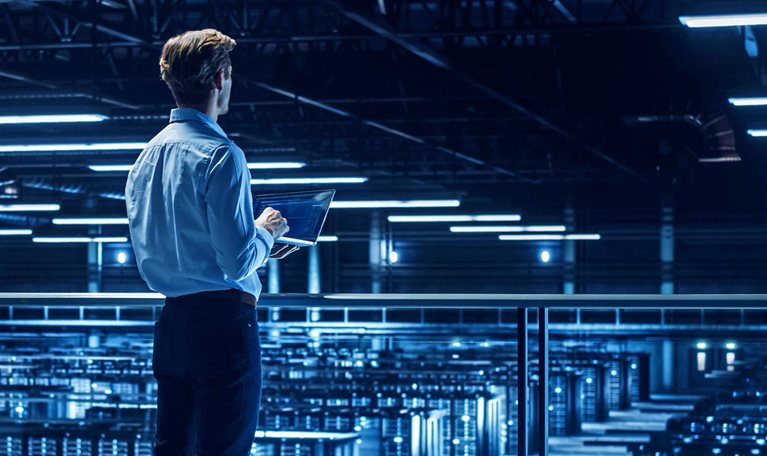 A person standing on an elevated walkway in a large, blue-lit data center, holding a laptop and overlooking rows of server racks.